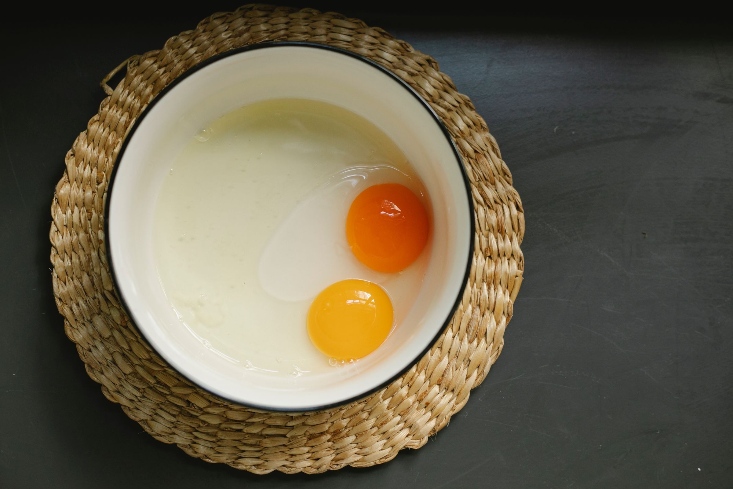 Two raw egg yolks in a white enamel bowl on a woven mat against a dark surface.