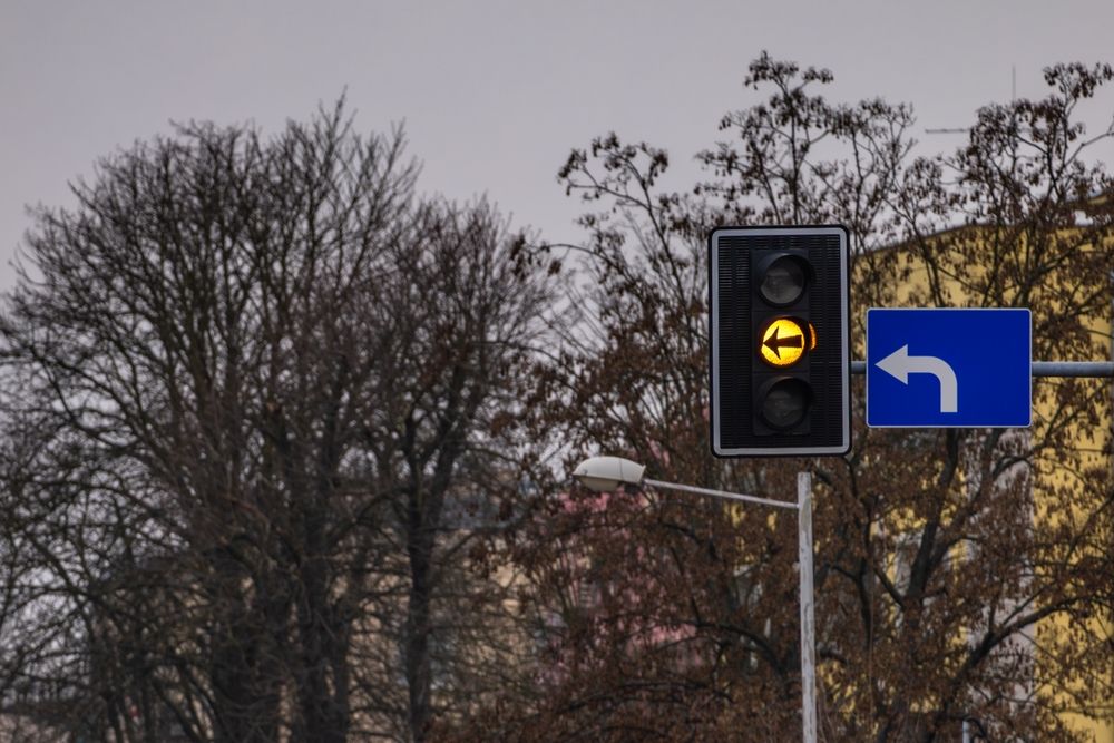 A yellow left-turn arrow traffic signal and blue directional sign set against bare winter trees, symbolizing the hazards of holiday shopping traffic.