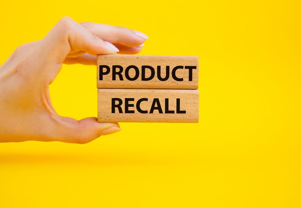 A hand holding wooden blocks that read "PRODUCT RECALL" against a yellow background, illustrating the Arrow International Recalls for Arrow FiberOptix and UltraFlex IAB Catheter Kits.