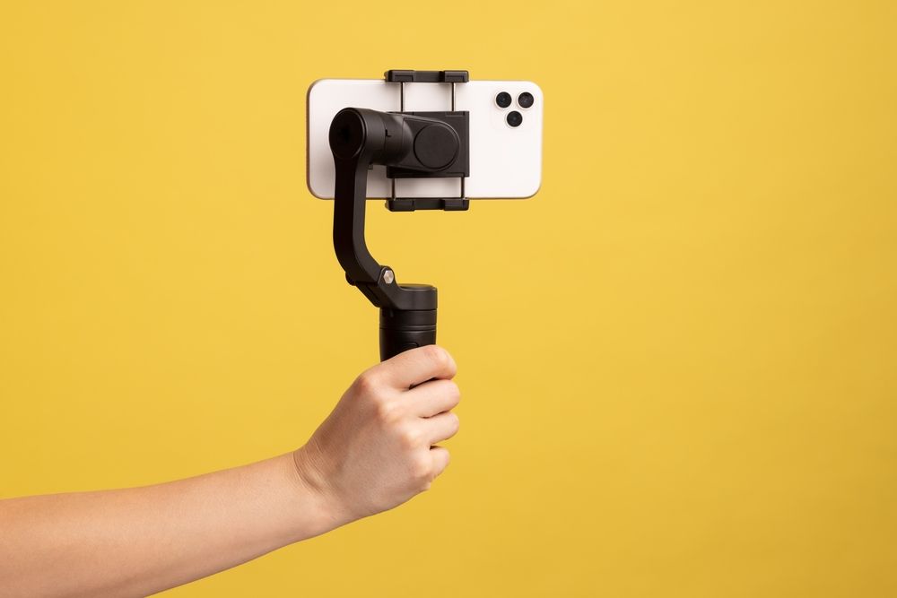 Hand holding a smartphone mounted on a stabilizing gimbal against a yellow background, representing livestreaming and online content creation.