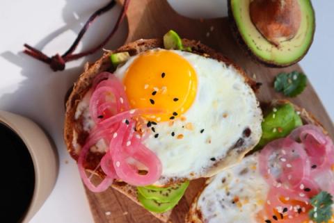 Avocado toast with homemade toasted sourdough bread, fried eggs, pickled red onion and cilantro on wooden board and coffee on the side.