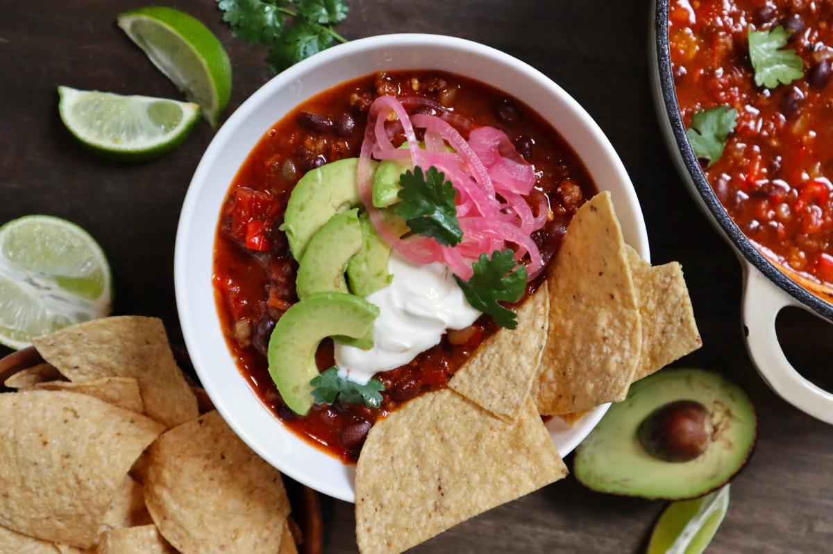 Overhead view of chili con carne served with tortilla chips, lime, avocado, and pickled onions