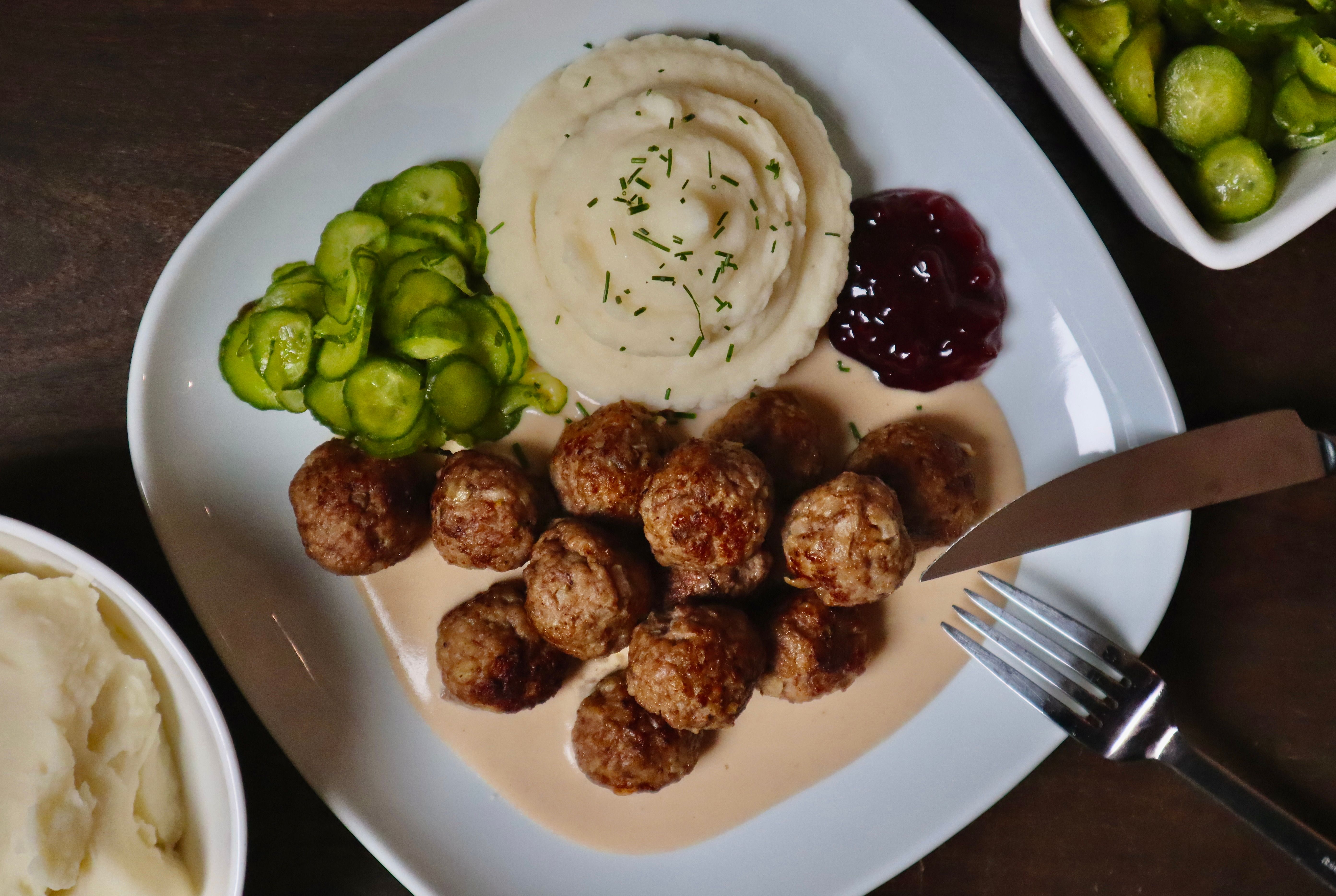 Swedish meatballs with creamy brown sauce, mashed potatoes, pickled cucumber and lingonberry jam on a white plate.