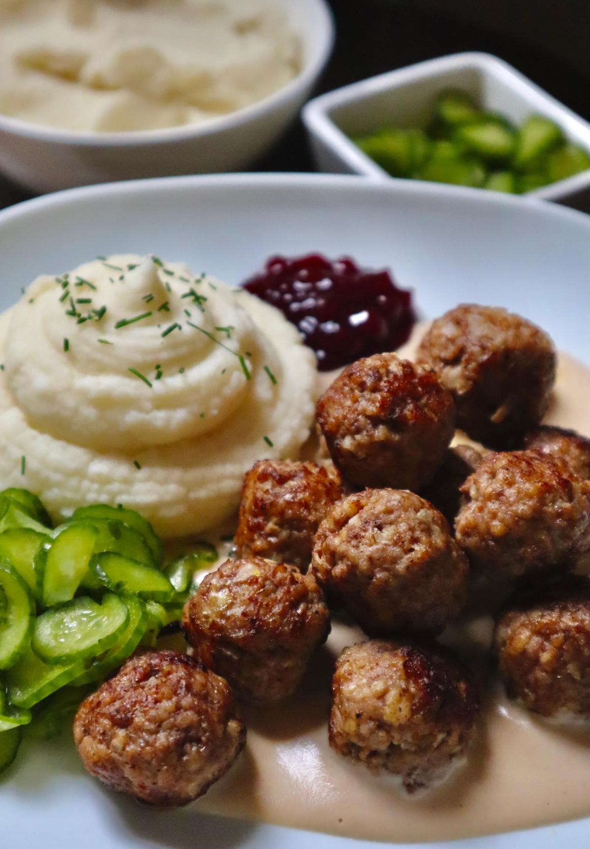 Close-up of homemade Swedish meatballs in creamy brown sauce with mashed potatoes, lingonberry jam and pickled cucumber.