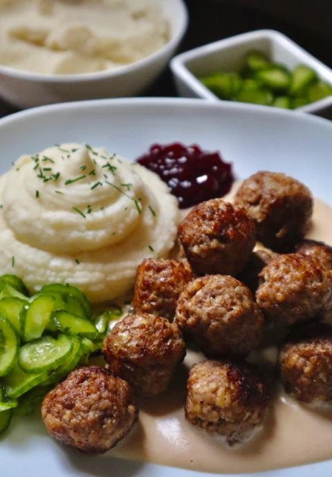 Close-up of homemade Swedish meatballs in creamy brown sauce with mashed potatoes, lingonberry jam and pickled cucumber.