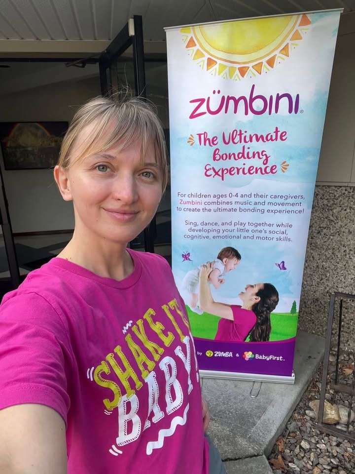Iryna Viriasova in a pink Zumbini shirt beside the Zumbini music and movement banner.