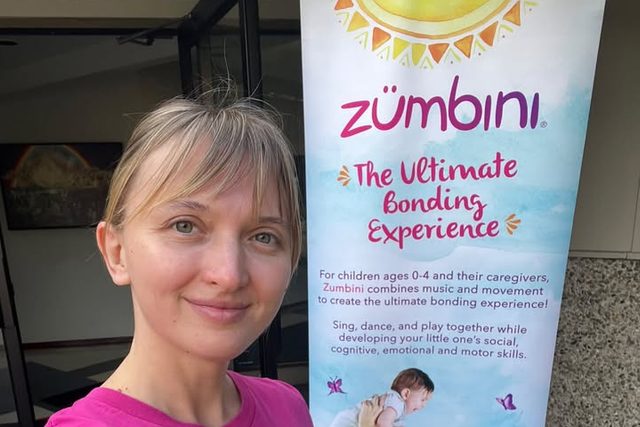 Iryna Viriasova in a pink Zumbini shirt beside the Zumbini music and movement banner.