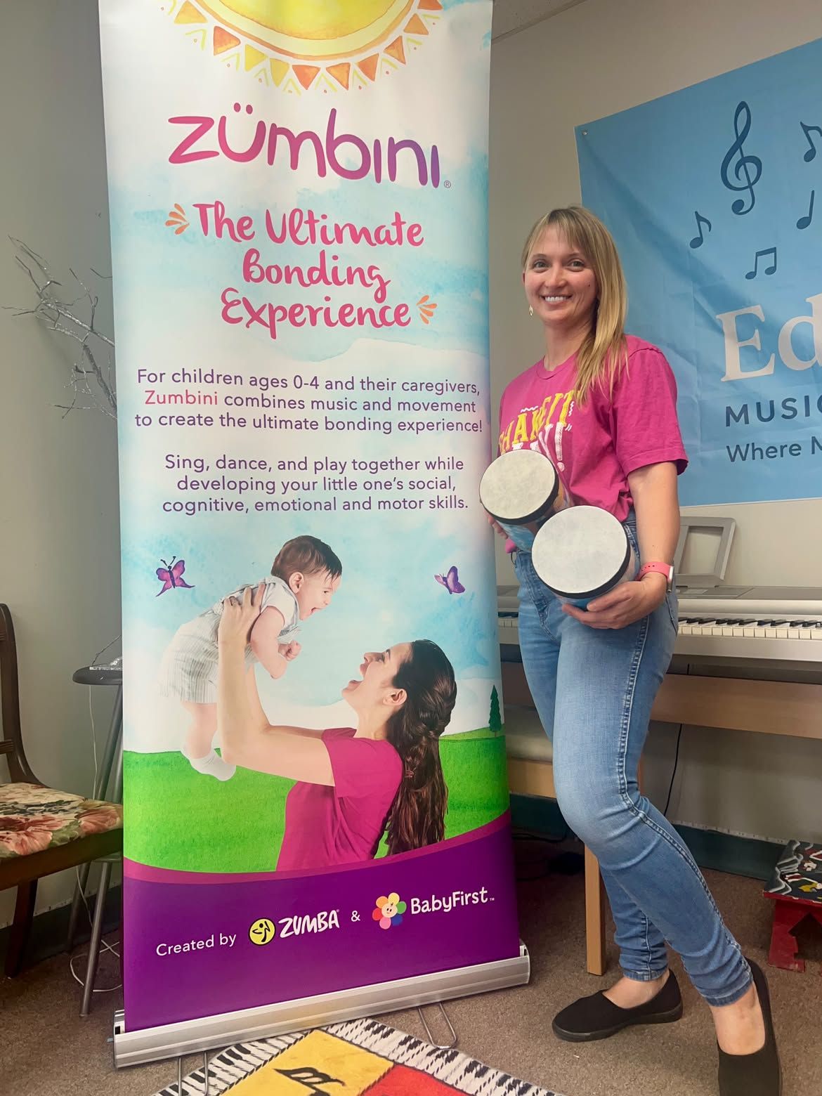 Iryna Viriasova holding hand drums beside a Zumbini banner at Edelweiss Music & Art Studio.