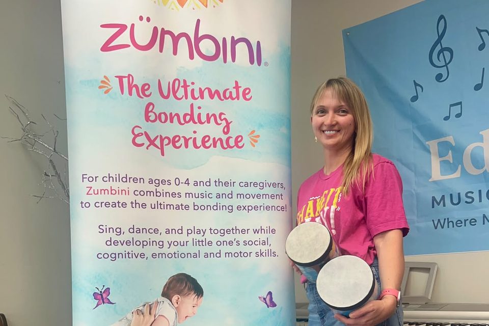 Iryna Viriasova holding hand drums beside a Zumbini banner at Edelweiss Music & Art Studio.