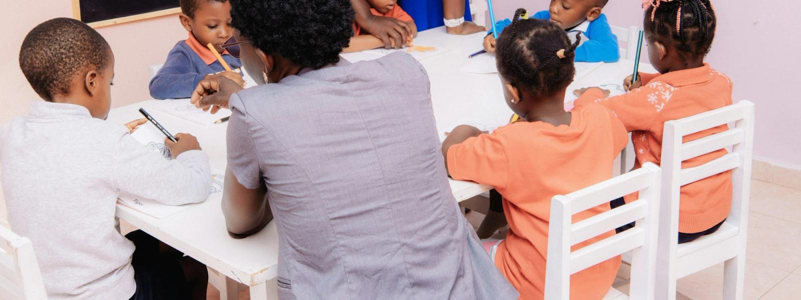 A teacher seated with a group of children drawing at a low table