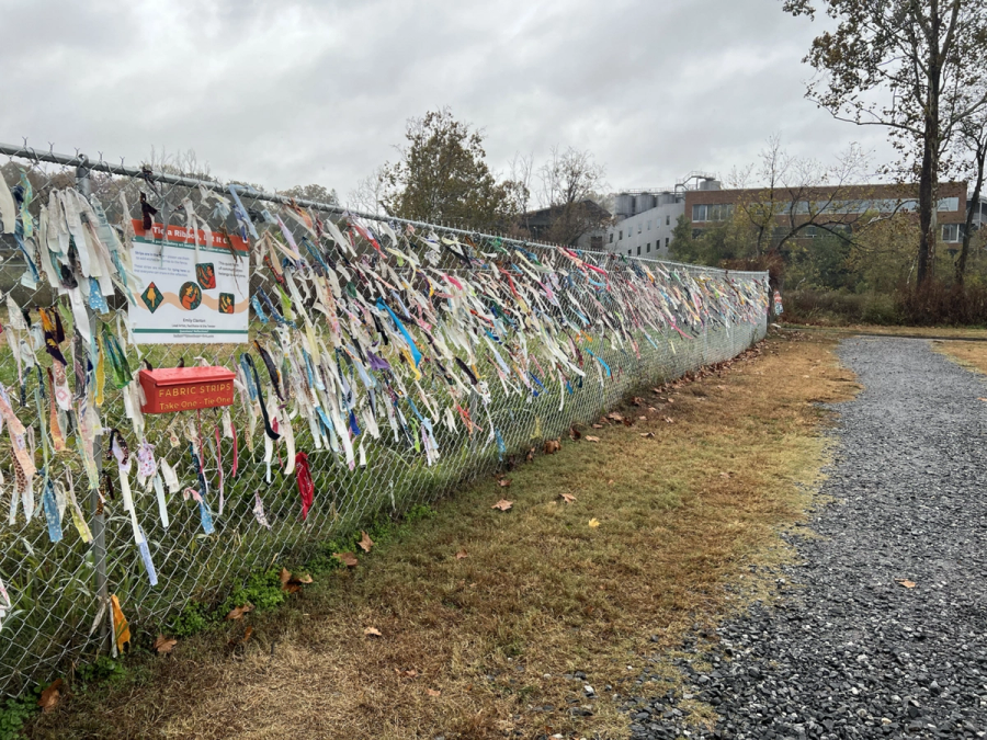 A wide autumn view of The Ties That Bind Asheville, a community-driven textile installation created by artist Emily Clanton in response to Hurricane Helene. The work invited the public to tie repurposed fabric strips onto a 81-foot chain-link fence along Riverside Drive in Asheville, North Carolina. This photograph, taken on 29 October 2025, shows the installation near the end of its active period, densely covered with contributions from the community. By Ecclantonart - Own work, CC BY-SA 4.0, https://commons.wikimedia.org/w/index.php?curid=178648722