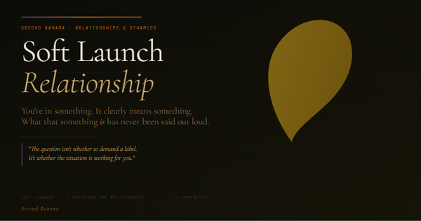 Hero image for “What Is a Soft Launch Relationship, and Is It Working For You?” Dark near-black background. A short purple-to-amber-to-terracotta rule sits above the eyebrow text “SECOND BANANA · RELATIONSHIPS & DYNAMICS.” The main headline reads “Soft Launch” in large light serif type, with “Relationship” below it in italic amber. Subhead: “You’re in something. It clearly means something. What that something is has never been said out loud.” A pull quote in italic reads: “The question isn’t whether to demand a label. It’s whether the situation is working for you.” Tags along the bottom: SOFT LAUNCH / DEFINING THE RELATIONSHIP / AMBIGUITY. A partially visible banana in gold tones appears in the lower right. Second Banana branding in amber italic.
