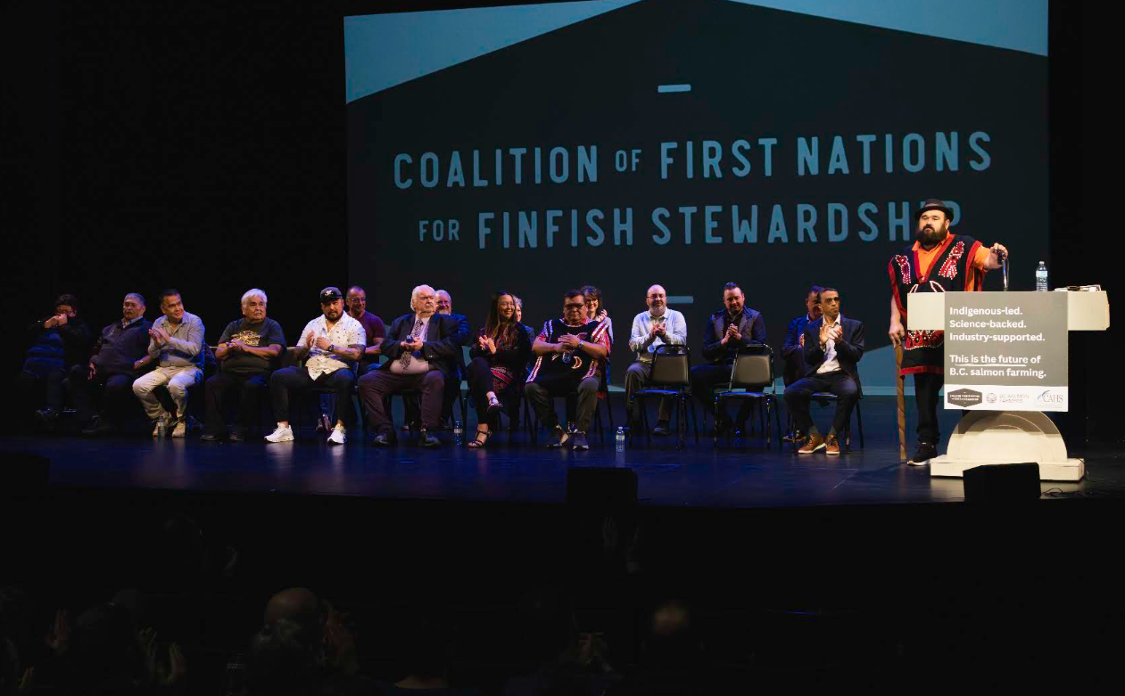A speaker in traditional regalia addresses an event from the Coalition of First Nations for Finfish Stewardship podium, with two others seated on stage
