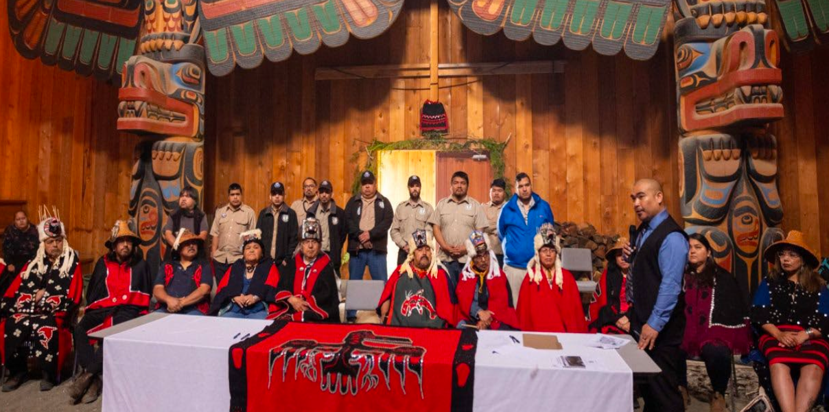 Hereditary chiefs in ceremonial regalia and Nation members gather at a signing table inside a carved big house on the B.C. coast