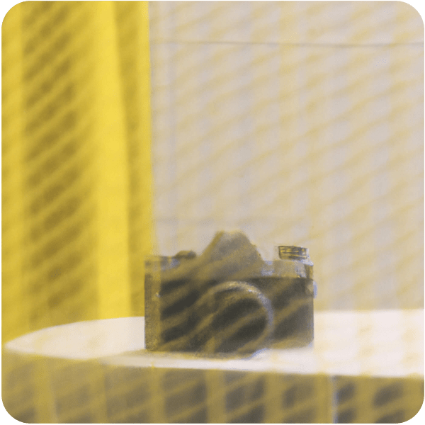 An analog style camera resting on a kitchen table in a calm, empty room seen through a net curtain