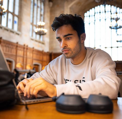 A young man is seated at a wooden table, actively typing on a laptop computer.