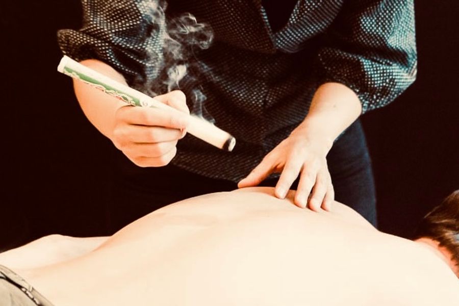 A practitioner holds a smoldering moxa stick a few inches above a patient's bare back, a thin plume of smoke rising from its tip, the other hand resting on the skin to judge the heat.