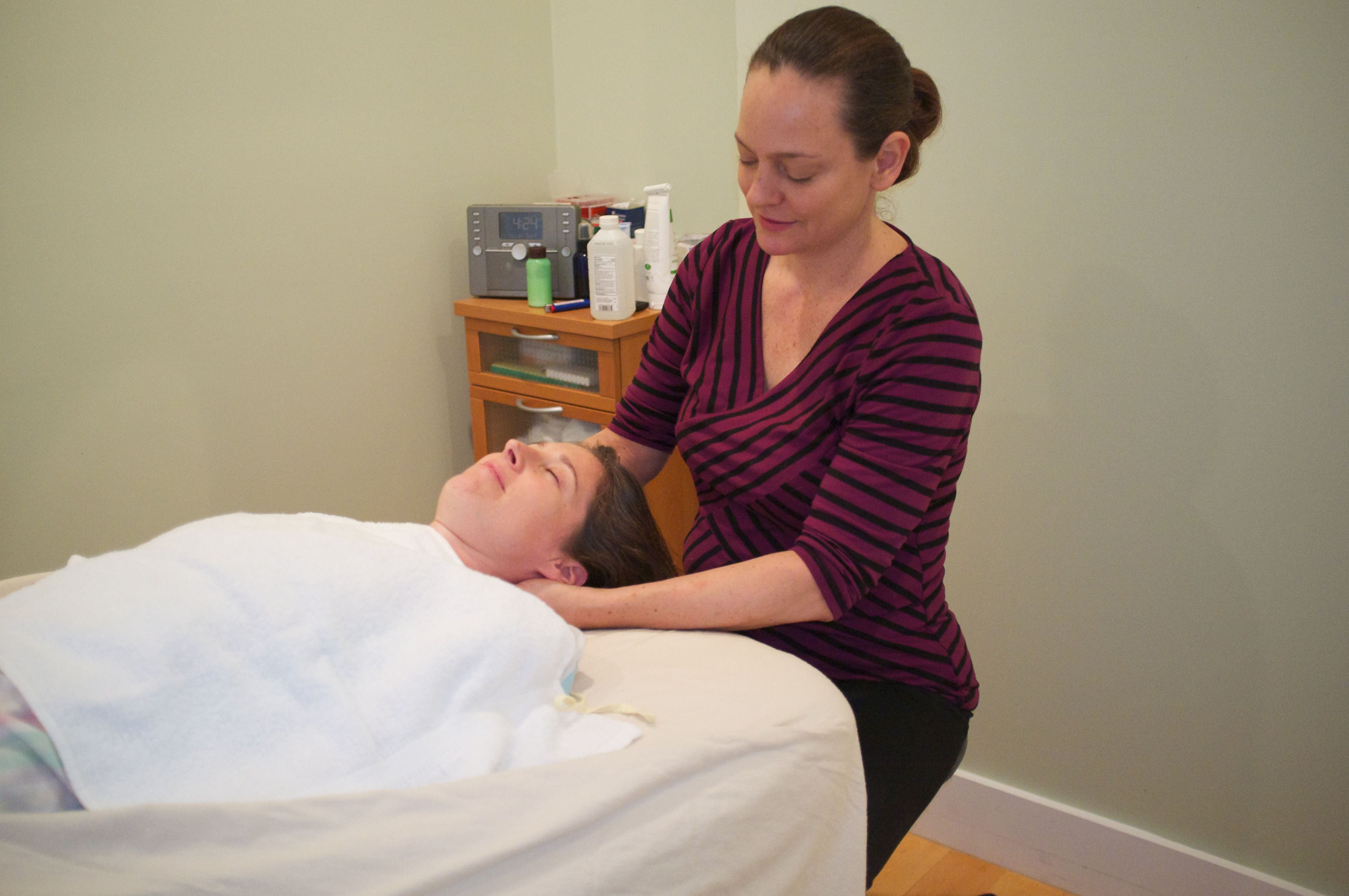 A practitioner stands at the head of the table with her hands cradling a patient's neck and jaw; the patient lies wrapped in a white towel with her eyes closed.