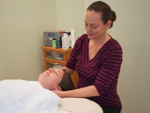 A practitioner stands at the head of the table with her hands cradling a patient's neck and jaw; the patient lies wrapped in a white towel with her eyes closed.