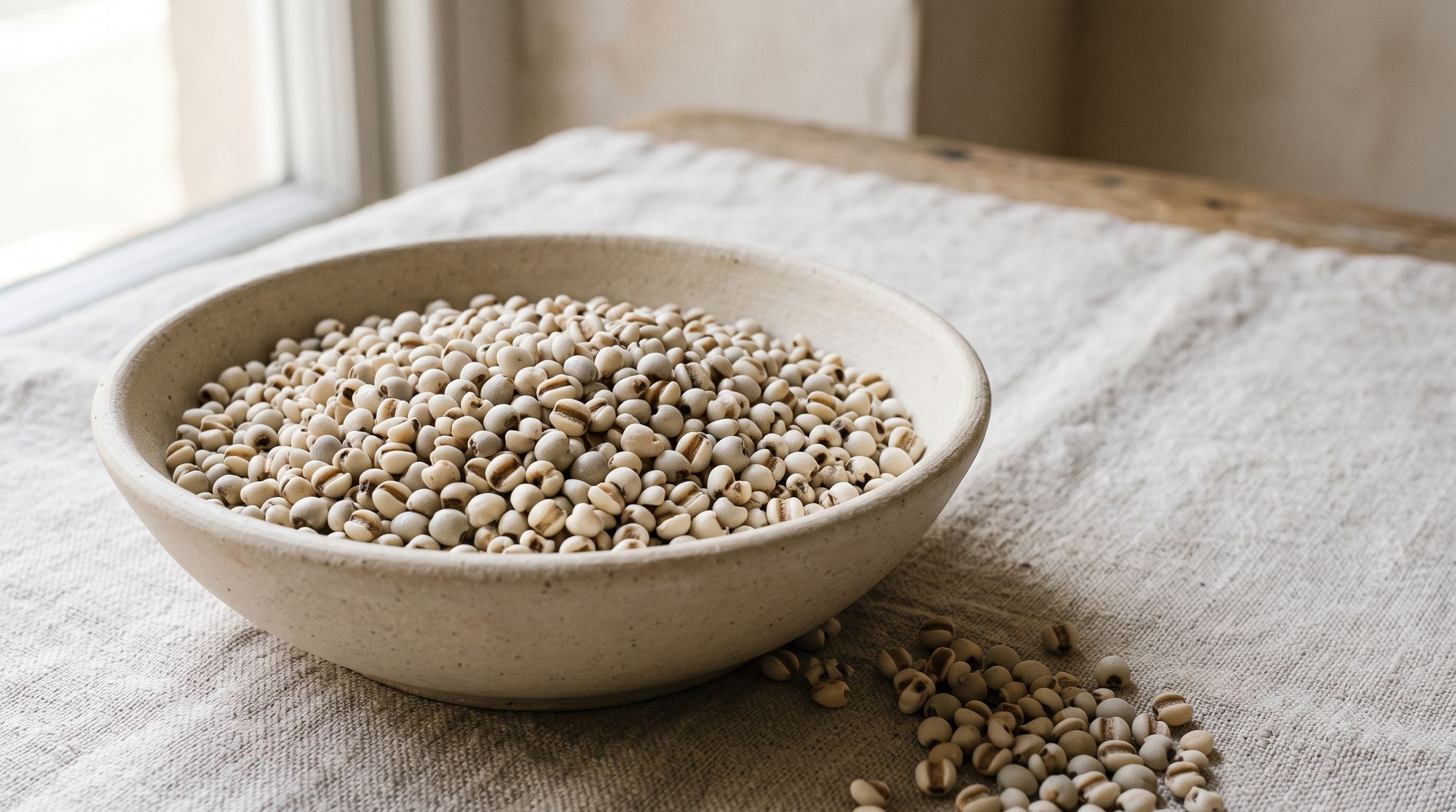 A bowl heaped with pale, rounded Job's tears, with more scattered on the linen tablecloth beside it.