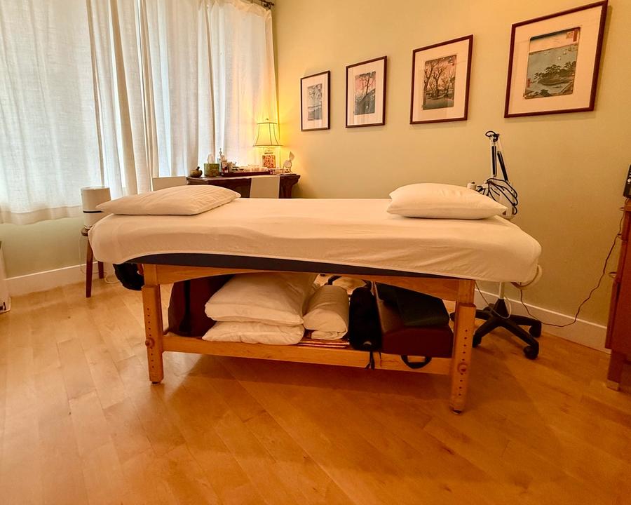 A treatment room with a made-up table under a curtained window, Japanese woodblock prints on the wall and a lamp lit on a side table.
