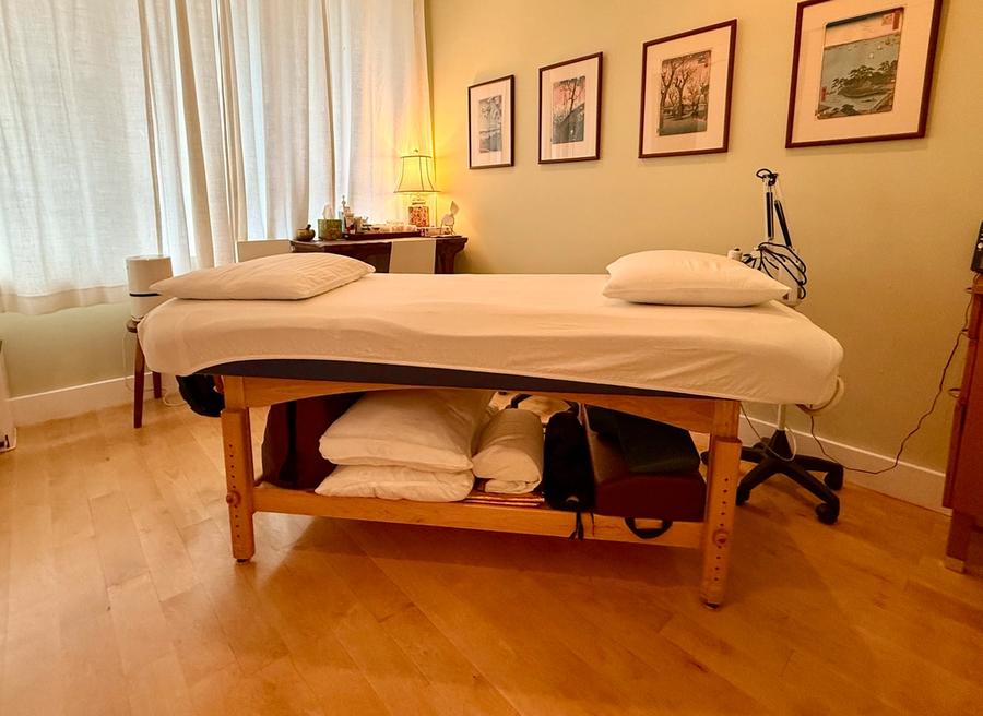 A treatment room with a made-up table under a curtained window, Japanese woodblock prints on the wall and a lamp lit on a side table.