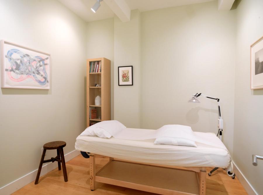 A small treatment room with a made-up table, a pale wood bookcase, an angled reading lamp and framed pictures on three walls; a wooden stool stands at the head of the table.