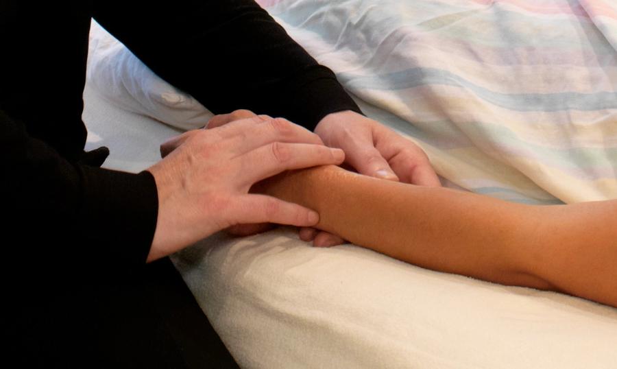 A practitioner's hands resting on a patient's forearm and wrist to take the pulse; the patient lies covered by a pastel-striped blanket.