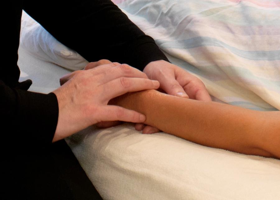A practitioner's hands resting on a patient's forearm and wrist to take the pulse; the patient lies covered by a pastel-striped blanket.