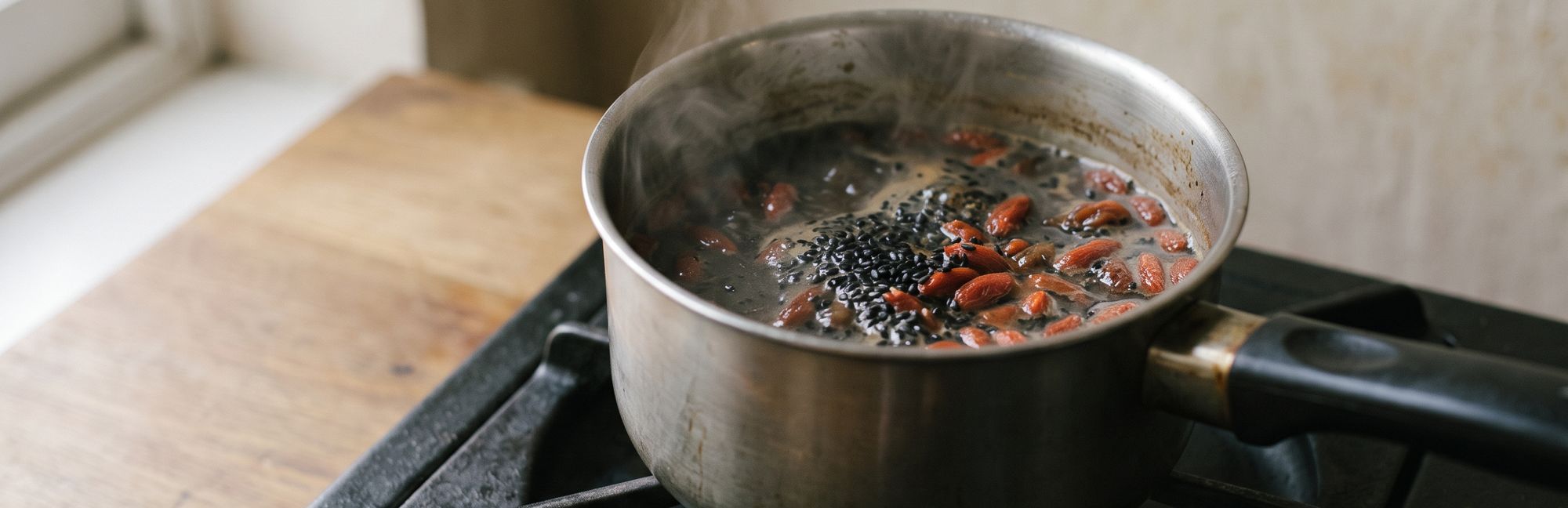 Black sesame seeds and goji berries simmering in a small steel pot on the stove, a thread of steam rising above it.