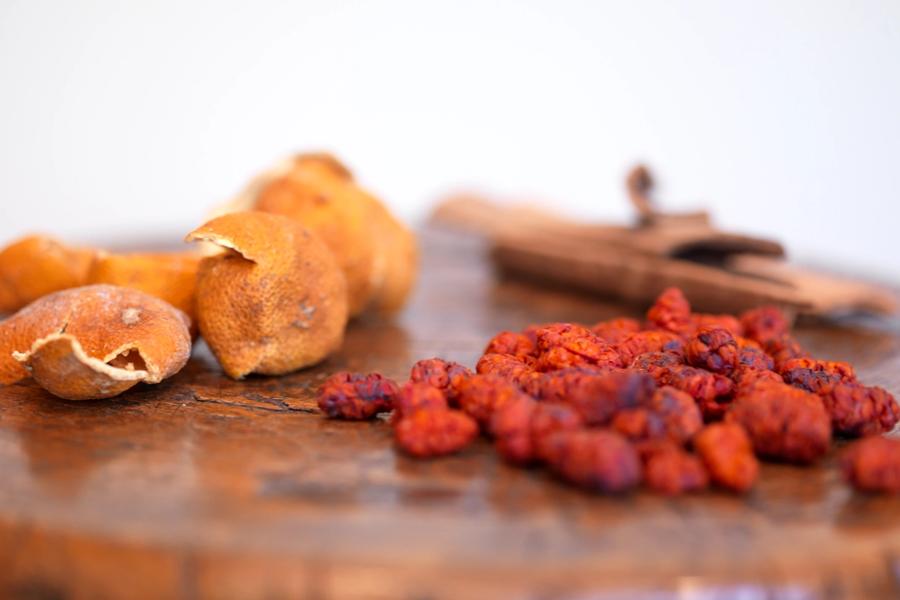 Dried tangerine peel, a cinnamon quill and a heap of dried red berries on a worn wooden board.
