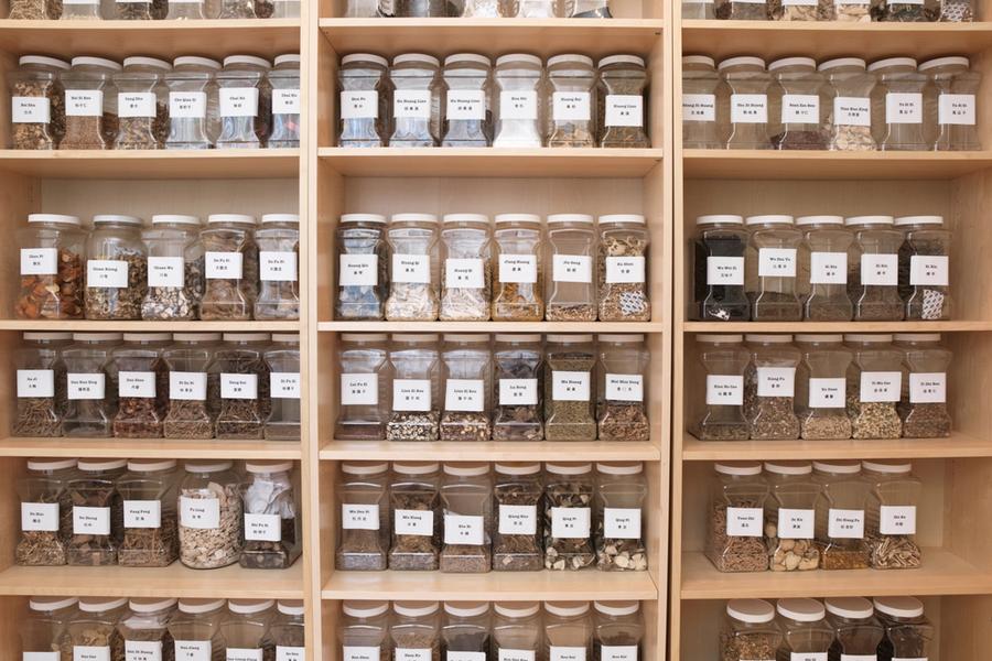 A wall of pale wood shelving filled with labeled glass jars of dried Chinese herbs, four rows deep, each jar named in English and Chinese.