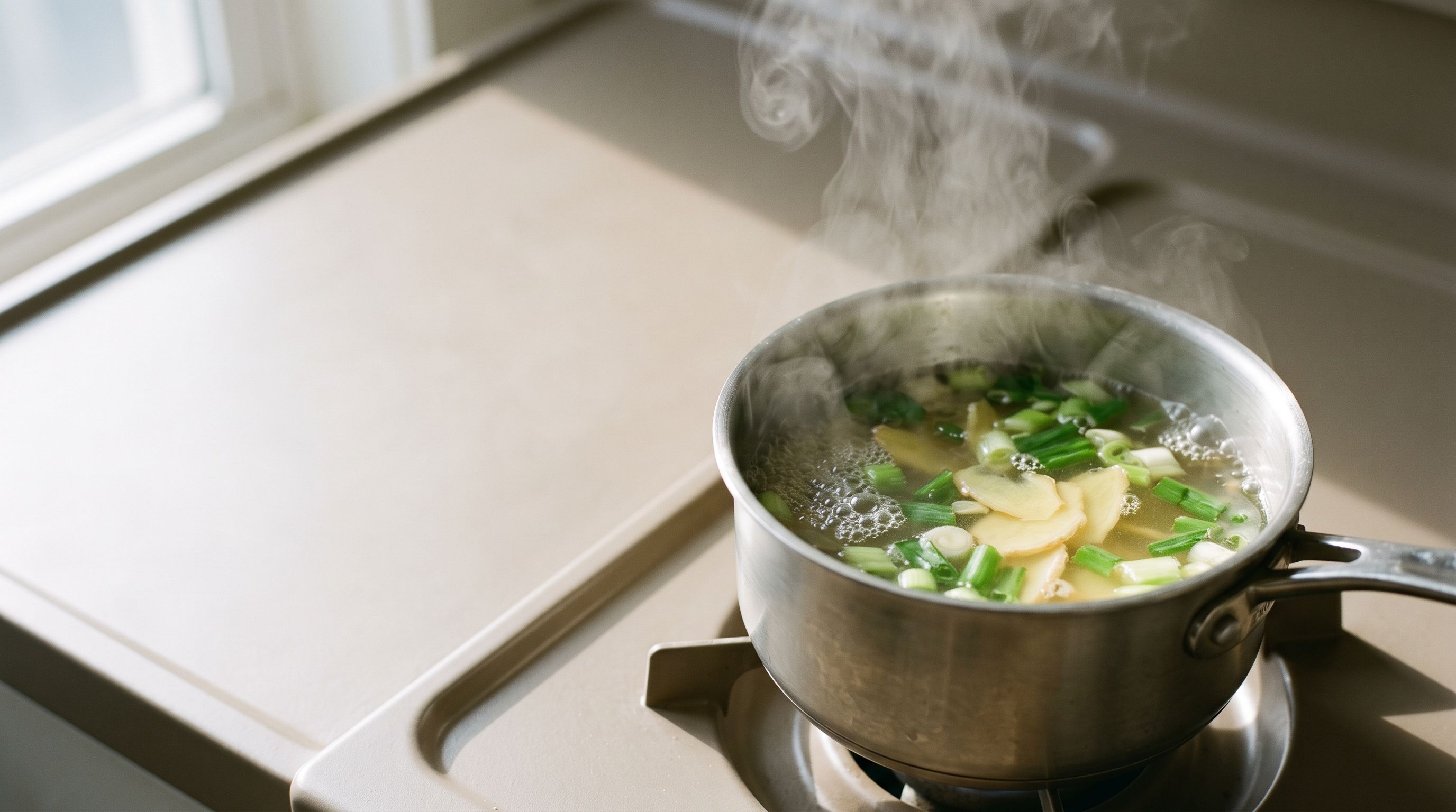 Ginger and scallion simmering in a small steel pot on the stove, steam rising in the window light.