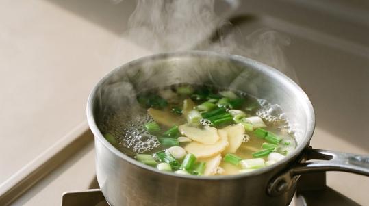 Ginger and scallion simmering in a small steel pot on the stove, steam rising in the window light.