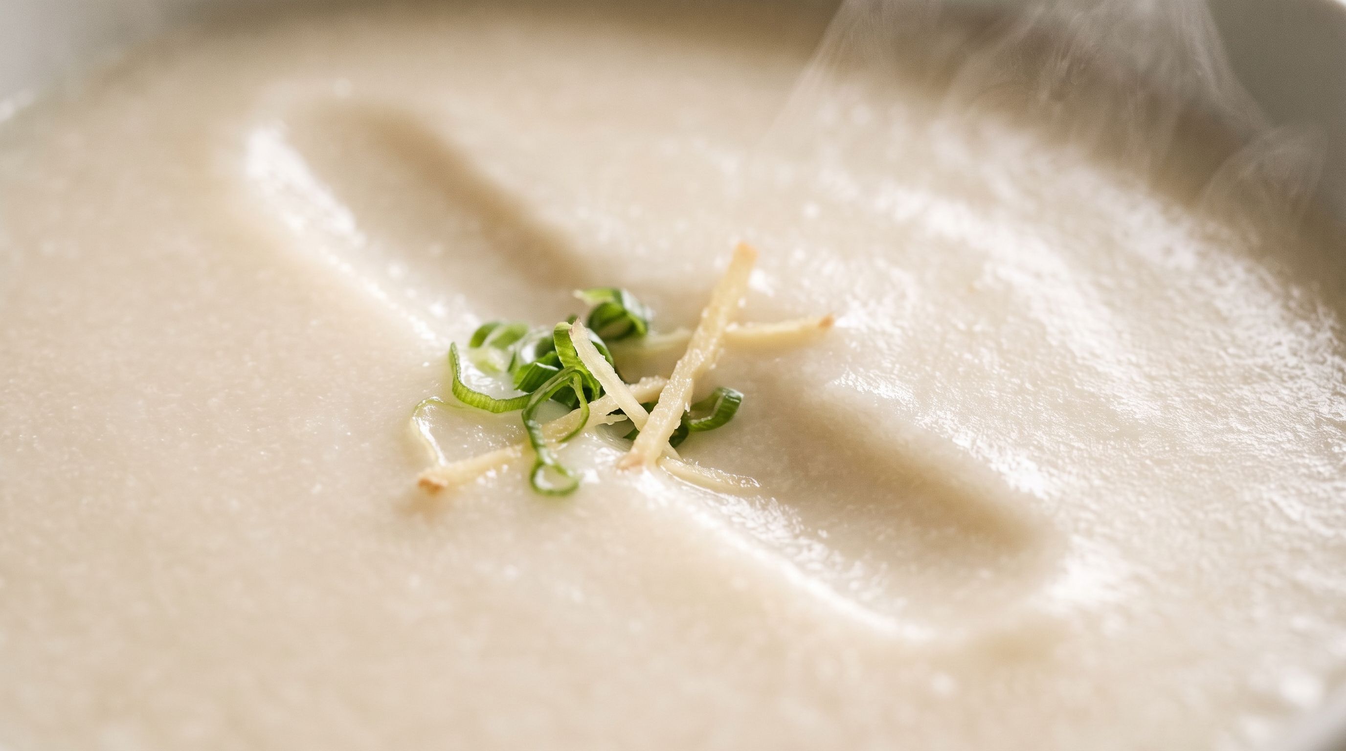 A close-up bowl of finished congee topped with shredded ginger and scallion, steam rising off the surface.