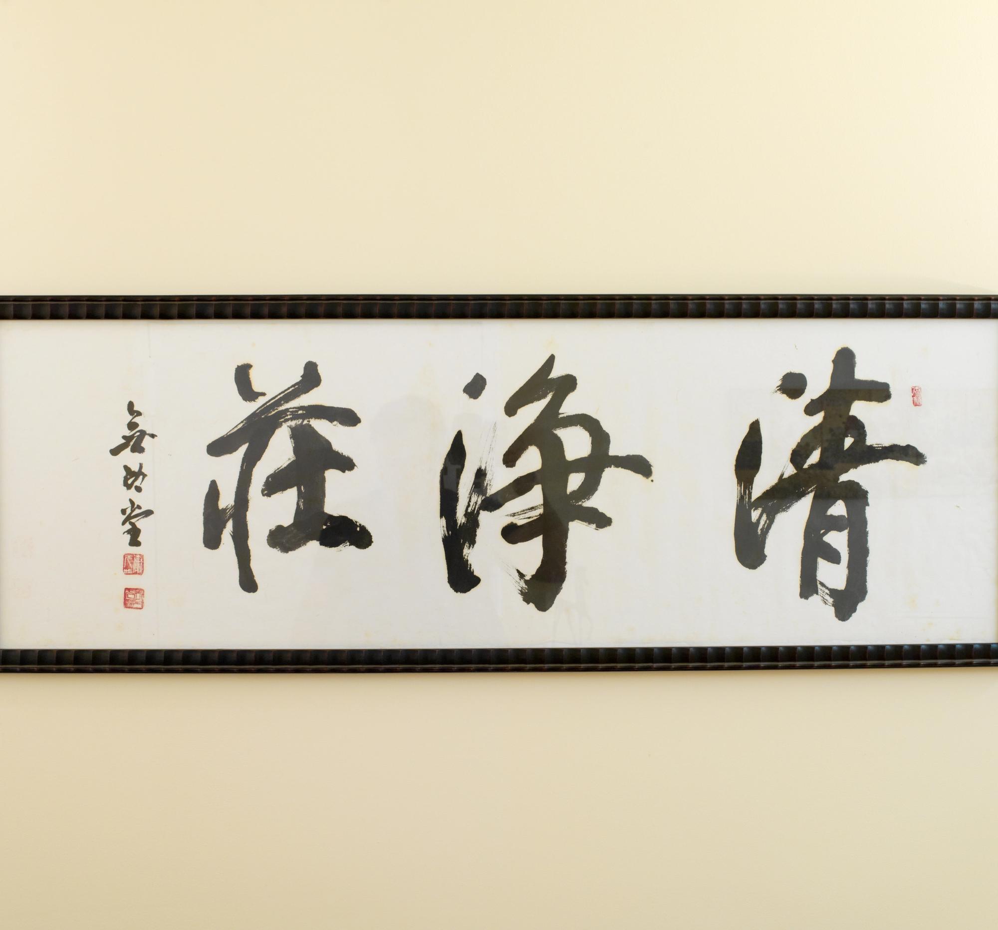 Framed brush calligraphy in black ink on white paper — three large characters with the artist's red seal — hung on a cream wall above the back of a bench.