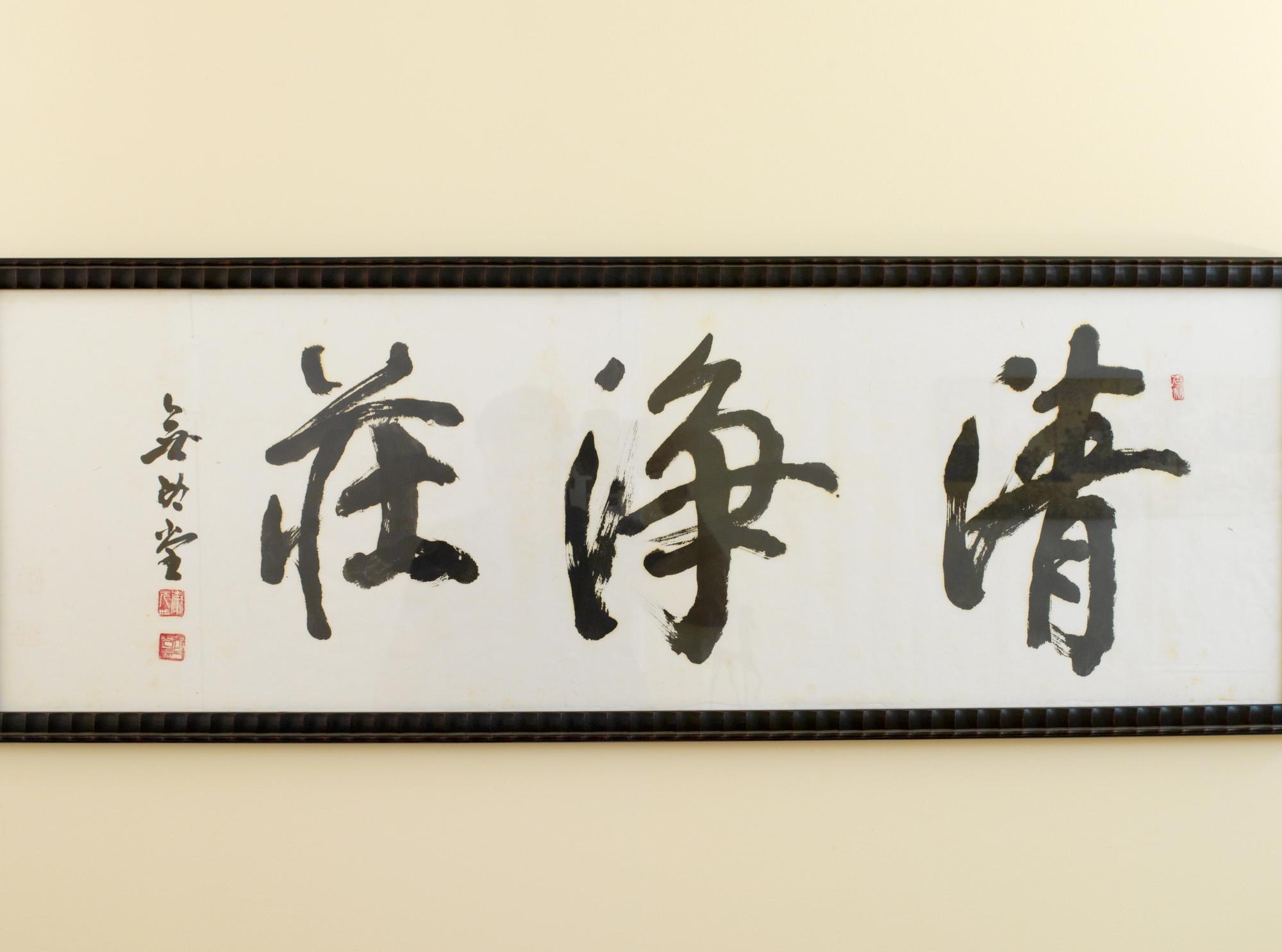 Framed brush calligraphy in black ink on white paper — three large characters with the artist's red seal — hung on a cream wall above the back of a bench.