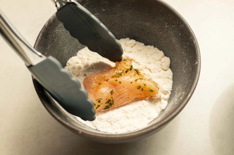 Tongs dredging chicken piece in a bowl of flour Tongs dredging chicken piece in a bowl of flour