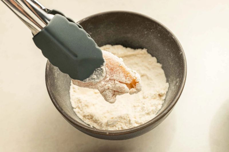 Tongs dredging chicken piece in a bowl of flour Tongs dredging chicken piece in a bowl of flour