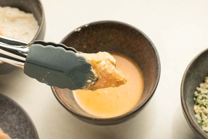 Tongs holding raw chicken piece coated in egg Tongs holding raw chicken piece coated in egg