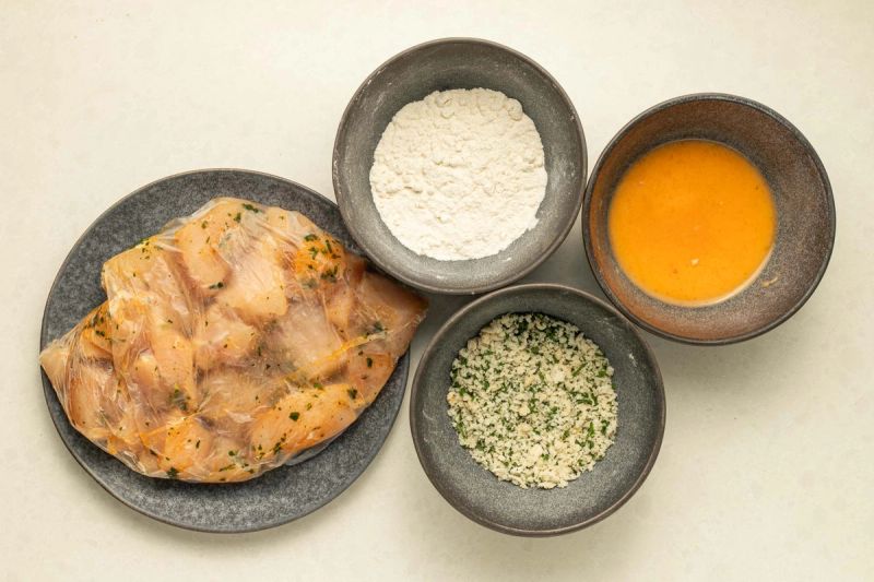 Ingredients spread of raw chicken, flour, egg, and breadcrumbs