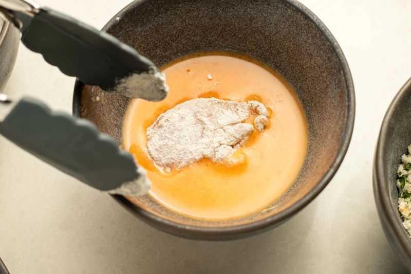 Tongs dropping chicken piece into a bowl of beaten egg