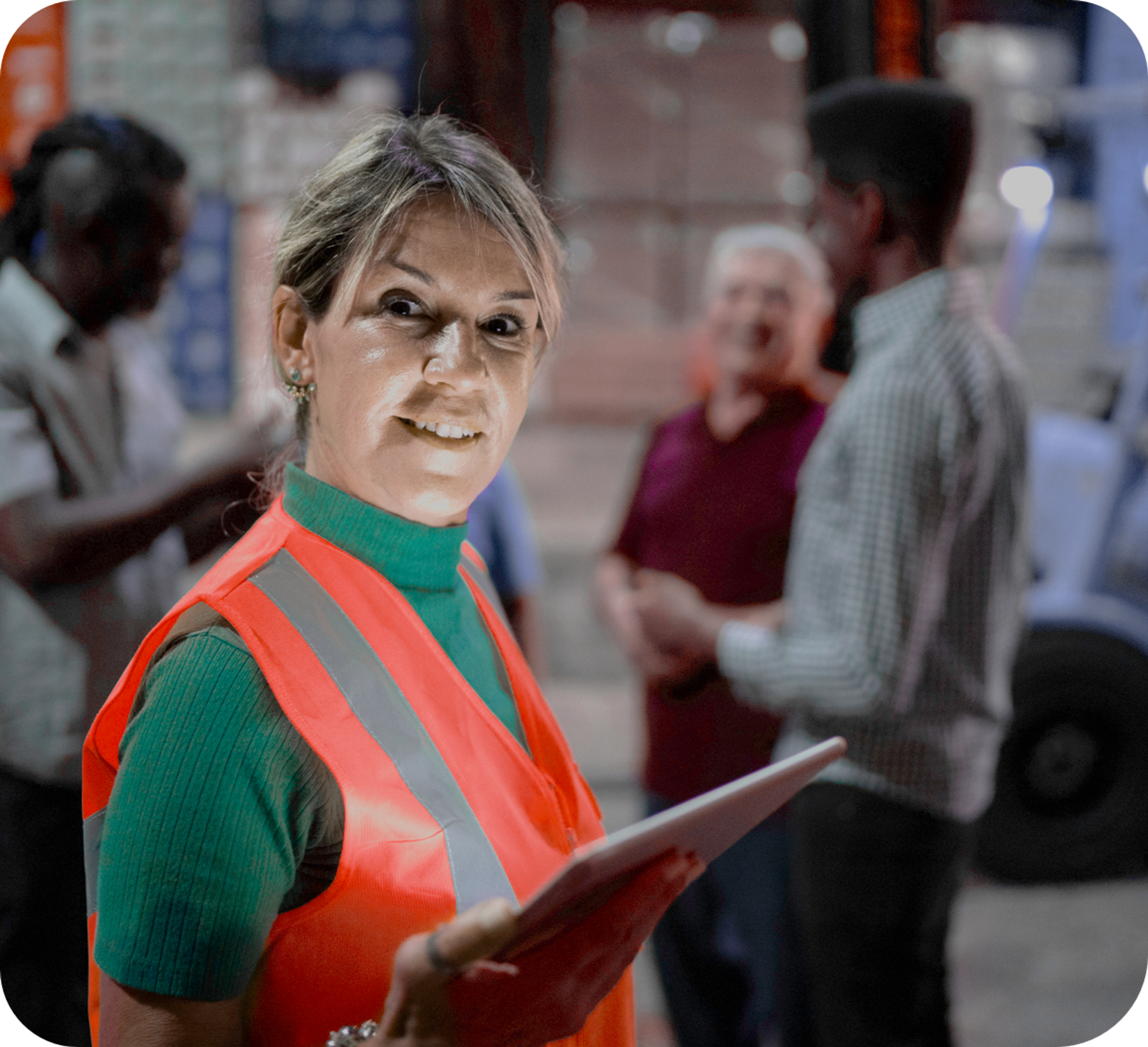 Femme d'âge moyen caucasienne dans un entrepôt portant un col roulé vert et une veste de sécurité orange, souriant et tenant un iPad, tandis que trois hommes sont flous en arrière-plan