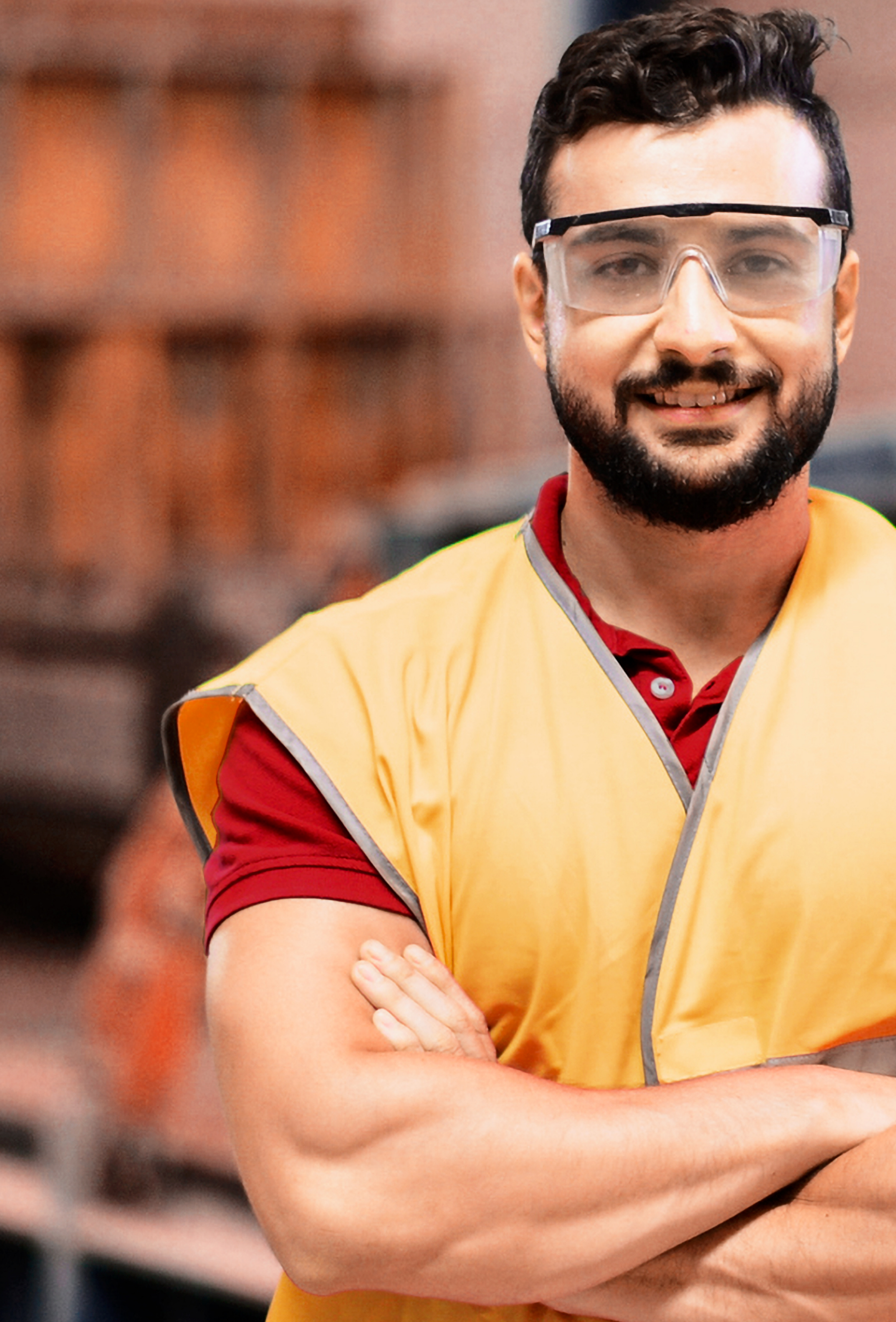 Picture of man with black moustache and beard wearing safety glasses, red shirt and orange safety vest inside a car battery warehouse