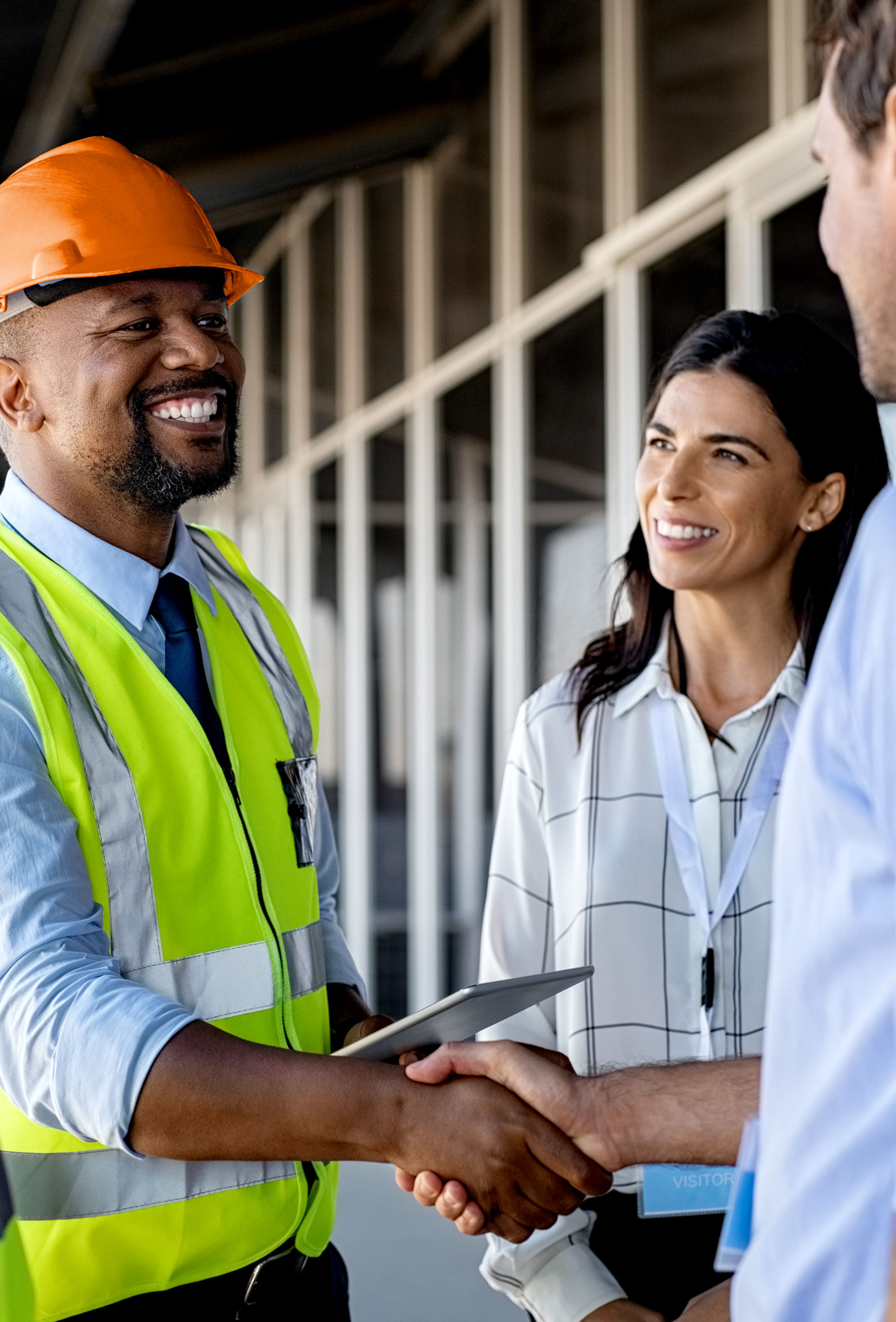 Picture of African man wearing an orange hard hat and yellow safety vest shaking hands with a Caucasian business man wearing a blue collared dress shirt, surrounded by a Caucasian woman smiling and wearing a white blouse and a Caucasian man wearing a dark blue dress shirt and yellow safety vest