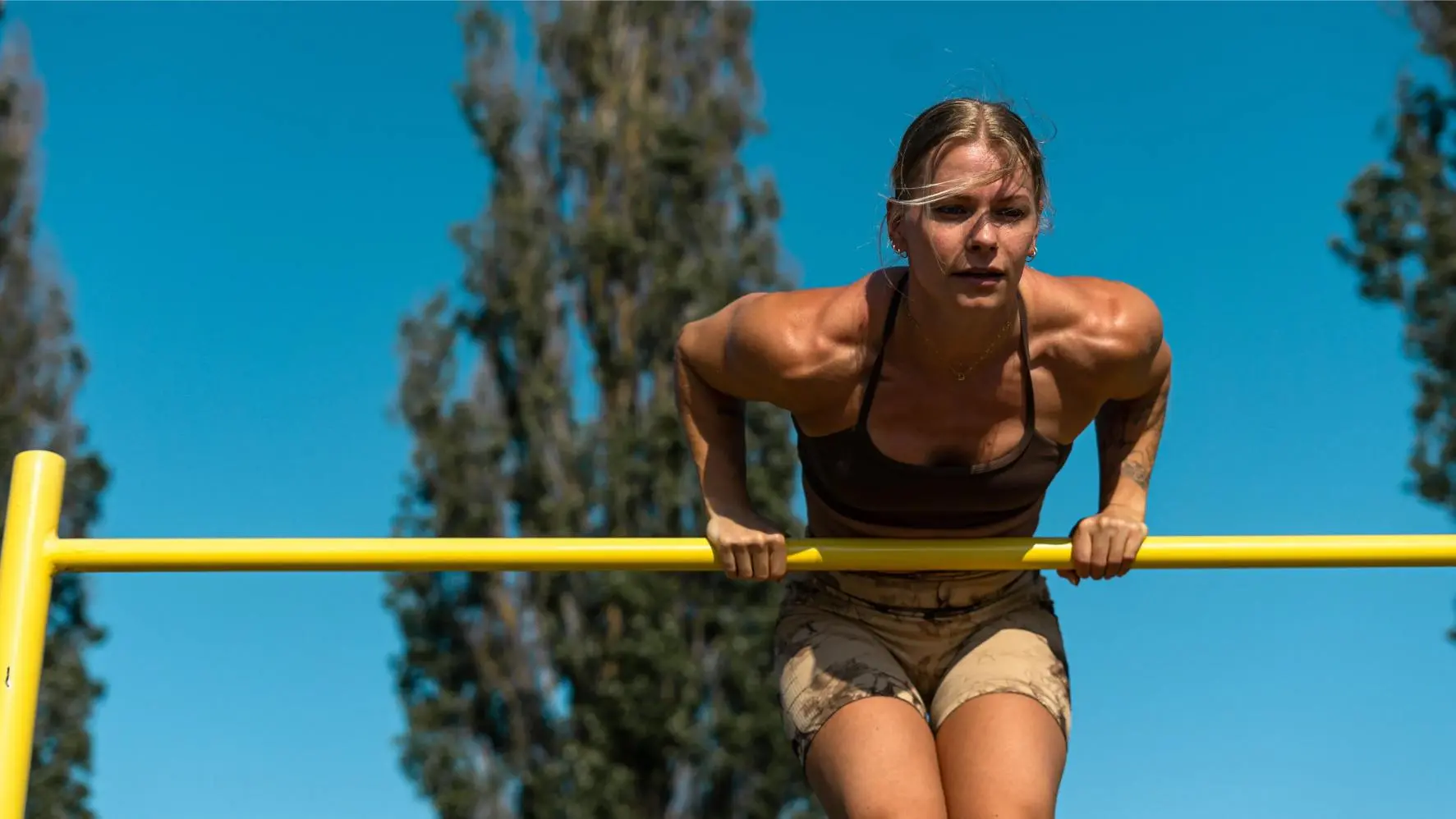 En kvinna utför en muscle-up på en gul stång mot en klar blå himmel.
