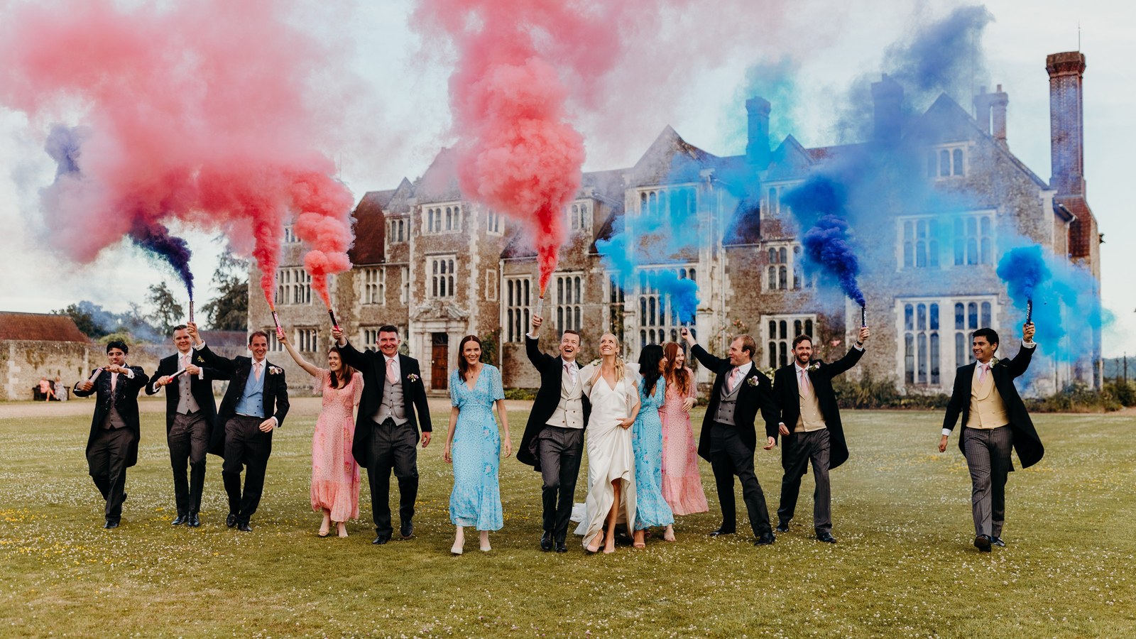 A group of wedding guests and the couple stand in a grassy field in front of a stately home