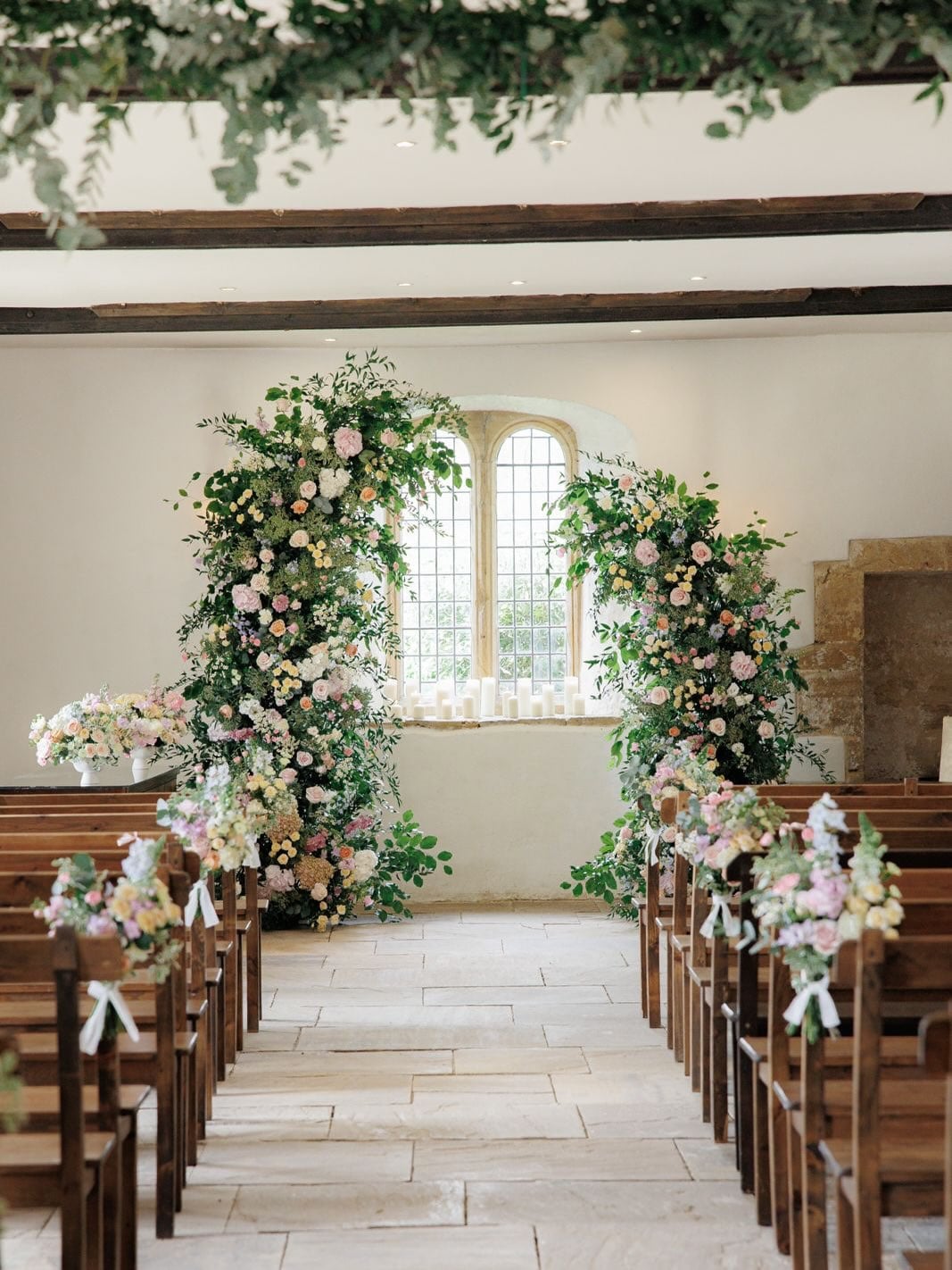 An aisle of wooden pews leads towards an ornate arch window, which is framed by abundant floral