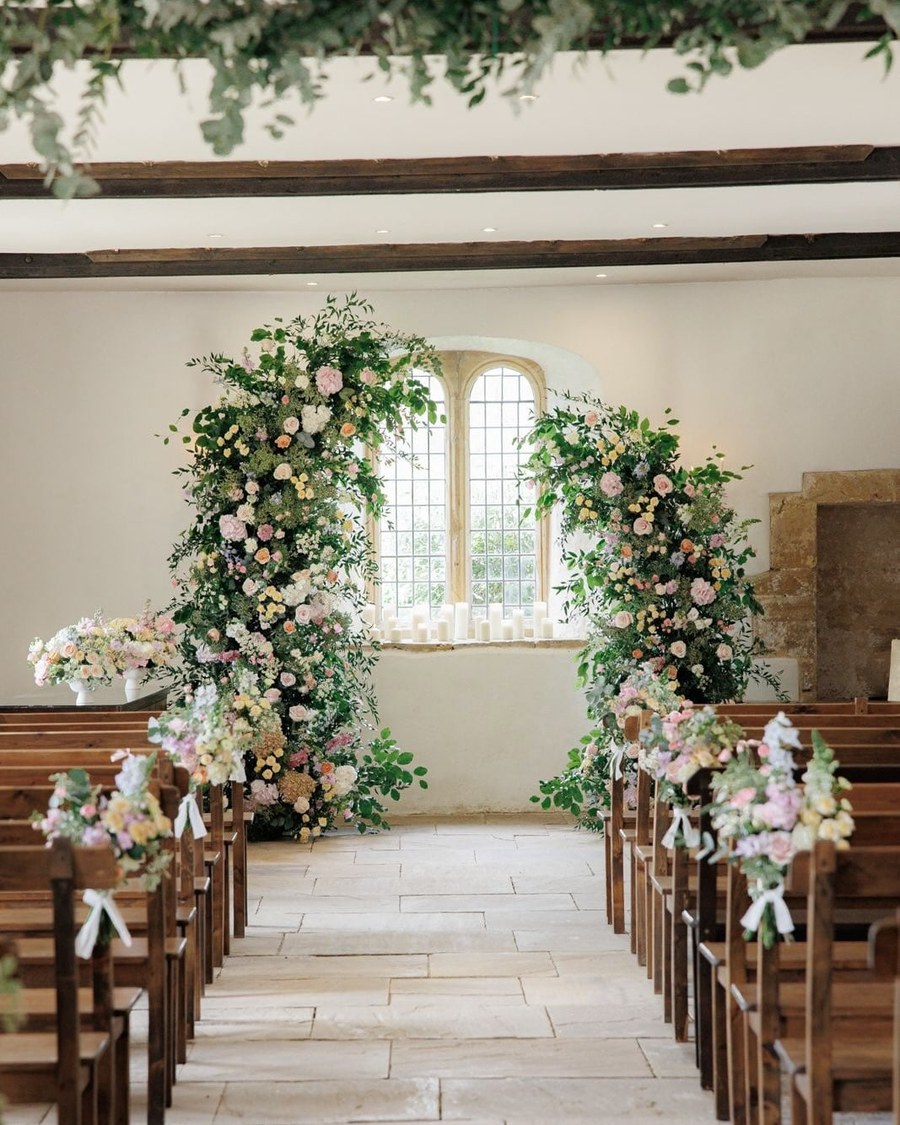 An aisle of wooden pews leads towards an ornate arch window, which is framed by abundant floral