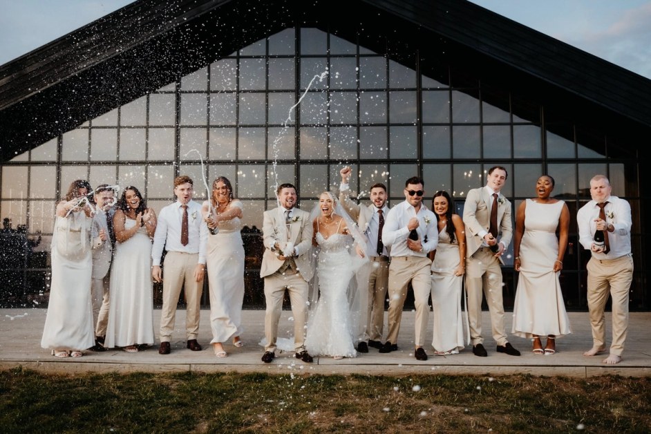 A group of wedding guests, including the bride and groom, stand in a line on a paved area outside a